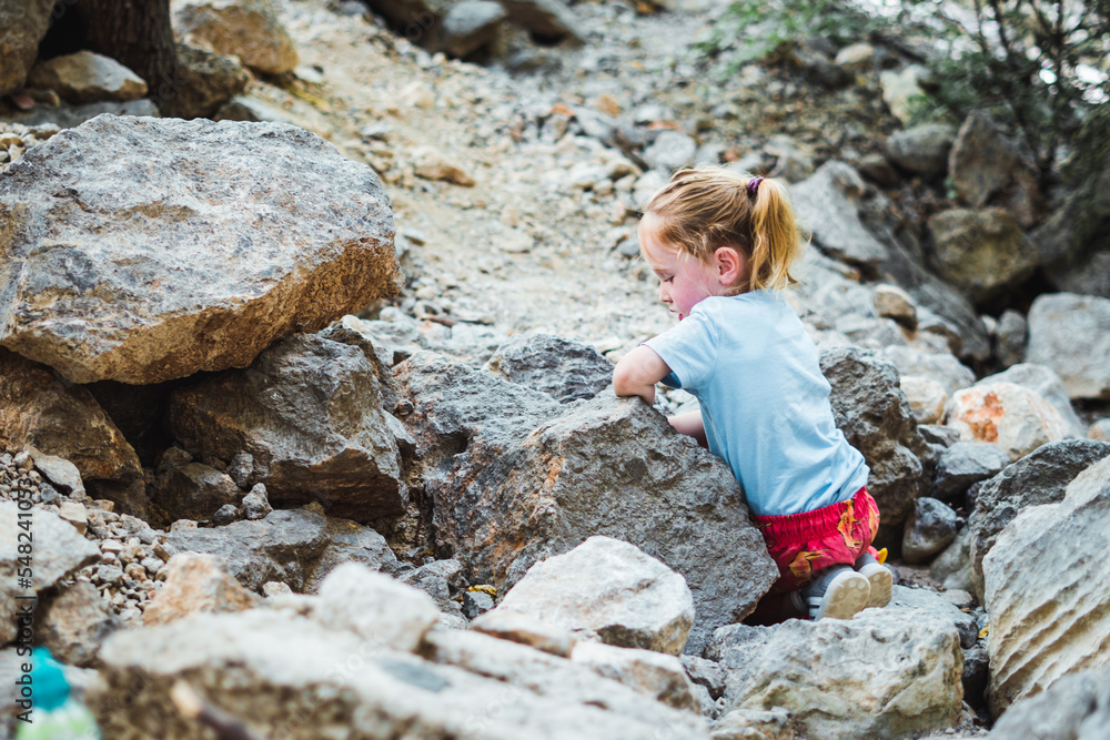 Small child rock climbing and independent play among rocks in a dry valley while on a hike in a natural park. Overcoming difficulty while developing motor skills in contact with nature.