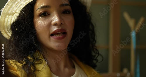 Handheld close up shot, Face of attractive young designer wearing stylish fashion hat smiling while using smartphone video call showing clothes during online sale in tailor shop