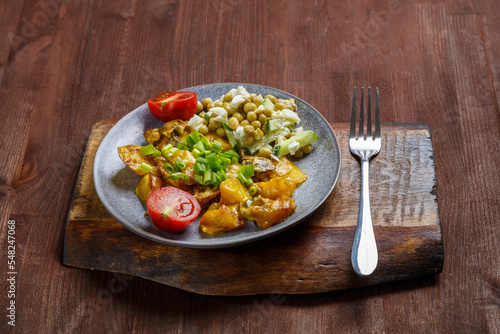 Baked potatoes with cheese and mushrooms garnished with tomatoes and peas sprinkled with green onions in a plate on a wooden board next to a fork.