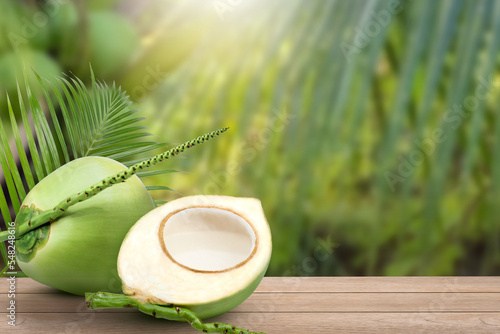 Young green coconut and half slice with juice on wooden table with coconut tree background.