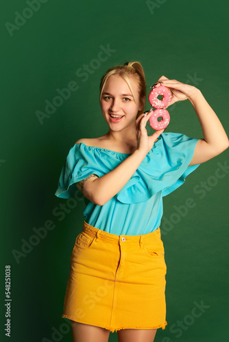 Emotional young beautiful girl, student in summer outfit is goofing around with glazed donuts isolated over green background. Positive emotions, happiness, hobbies and joy