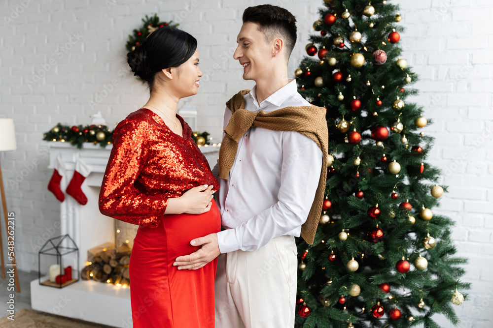 Obraz premium side view of pregnant and elegant asian woman with husband smiling at each other near Christmas tree in living room
