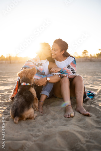 Hispanic lesbian couple sit on the beach and kiss with a blanket and dog