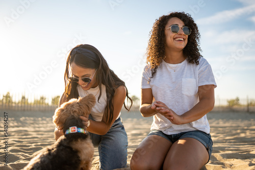 Hispanic lesbian couple sit on beach and pet dog