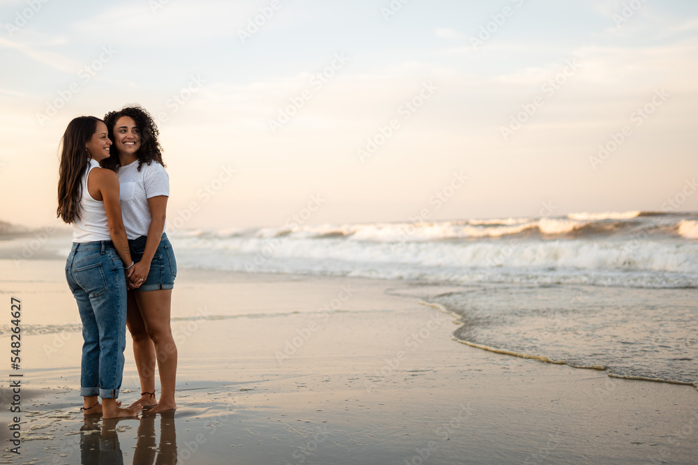 Hispanic lesbian couple hold hands at the beach while standing in wet sand