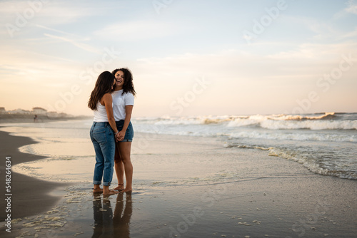 Hispanic lesbian couple hold hands at the beach while standing in wet sand