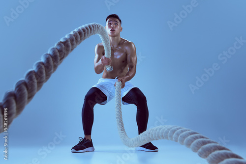 Portrait of young muscular man training, pulling rope isolated over blue background in neon light. Stength and power photo