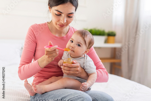 Adorable little baby girl eating from spoon, young caring mother feeding fer cute infant with mash fruit puree