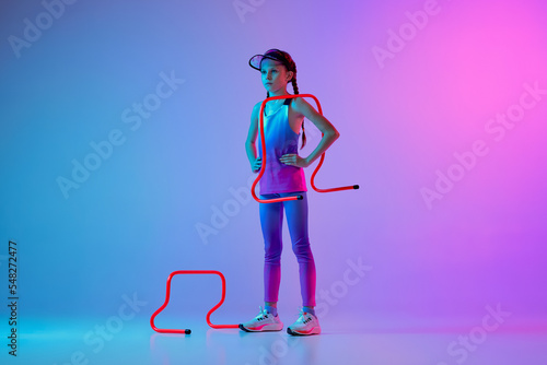 Motivation. Serious sportive girl, junior runner in stylish sportswear and cap posing isolated on gradient pink-blue background with neon filter, light.