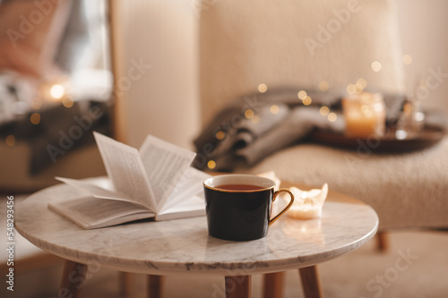 Canvas Print Cup of tea with paper open book and burning scented candles on marble table over cozy chair and glowing lights in bedroom closeup