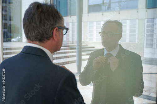 manager con giacca e cravatta  si riflette in una vetrata e si rimette in ordine in contesto urbano moderno photo