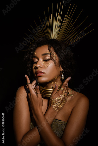 confident african american woman with golden necklace and paint on cheeks posing isolated on black.