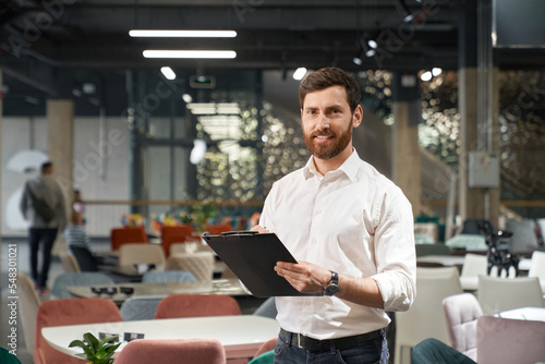 Successful entrepreneur in wearing shirt making notes, while working in modern expo center. Portrait of handsome restaurateur smiling at camera, while choosing furniture in store. Concept of business. photo