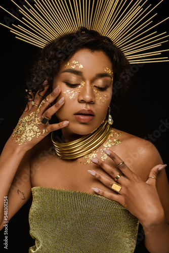 young african american woman with golden accessories and paint on face posing isolated on black.