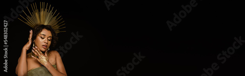 young african american woman with golden accessories and paint on face posing isolated on black, banner.
