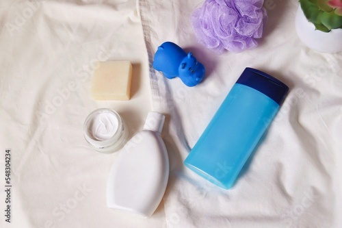 White shampoo bottle, blue shower gel package, facial cream jar, natural soap, purple sponge and rubber toy hippo. Hair and skin care cosmetics flat lay photo