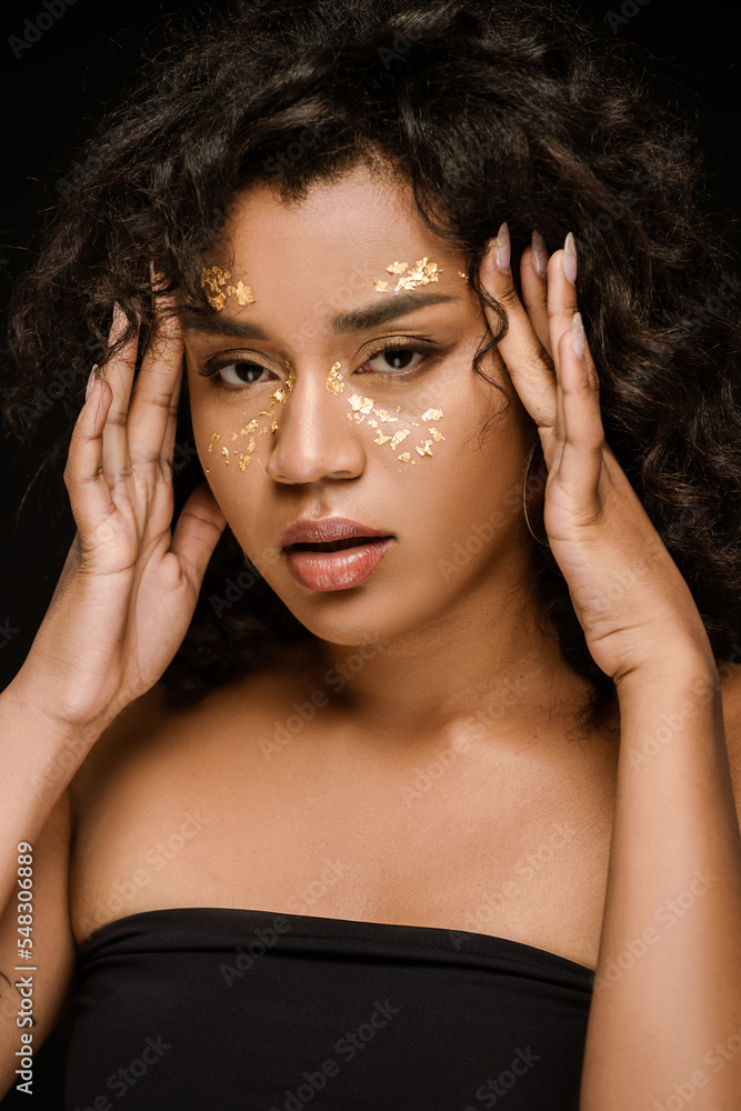 curly african american woman with golden paint on cheeks looking at camera isolated on black. curly african american woman with golden paint on cheeks looking at camera isolated on black.