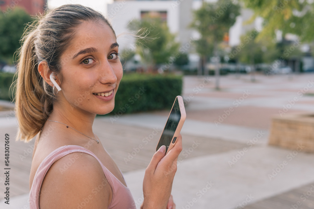 Sporty young woman with wireless earphones smiling at camera while holding her mobile phone to send an audio message. Sporty young woman with wireless earphones smiling at camera while holding her mobile phone to send an audio message.