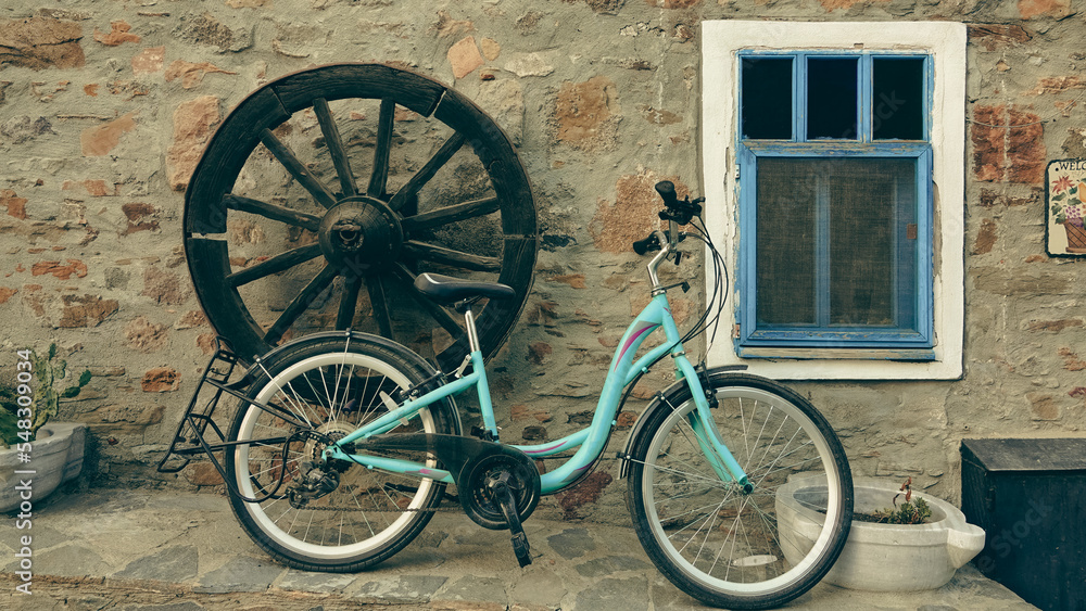 A bicycle in front of an old stone house with a window, a carriage wheel mounted decoratively on the wall, a flower pot for flowers made of marble on the floor