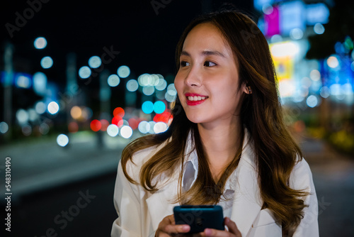 Business woman using mobile phone walking through night city street while waiting car to pick up home, Beautiful young smiling female texting work message on smartphone outside office, social media