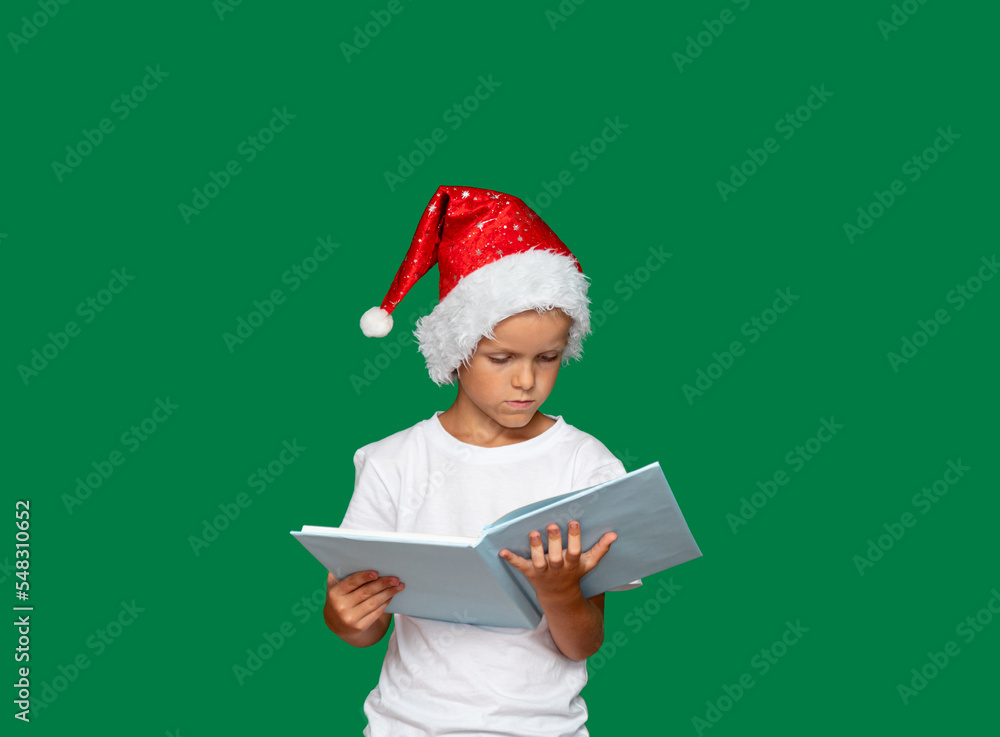 Boy in Santa Claus hat is thinking and looking at an open book, which he holds with his hands. Green background with space for text. Selective focus. Boy in Santa Claus hat is thinking and looking at an open book, which he holds with his hands. Green background with space for text. Selective focus.