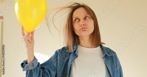 Young beautiful woman having fun with yellow air balloon indoors. Surprised girl is rubbing a balloon against her hair and electricity is generated. Laws of physics.
