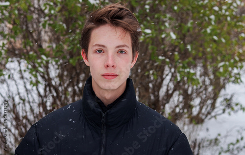 Portrait of a teenage boy close-up against the background of snow and a green bush.