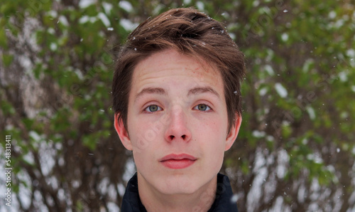 Portrait of a teenage boy close-up against the background of snow and a green bush.