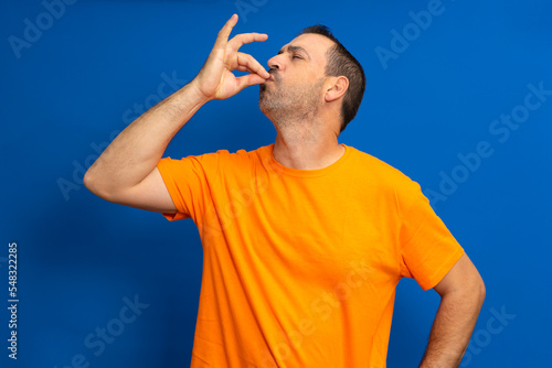 Portrait of a bearded hispanic man in orange t-shirt with puckered lips showing gourmet sign with fingers tasty delicious isolated on vibrant blue color background. photo