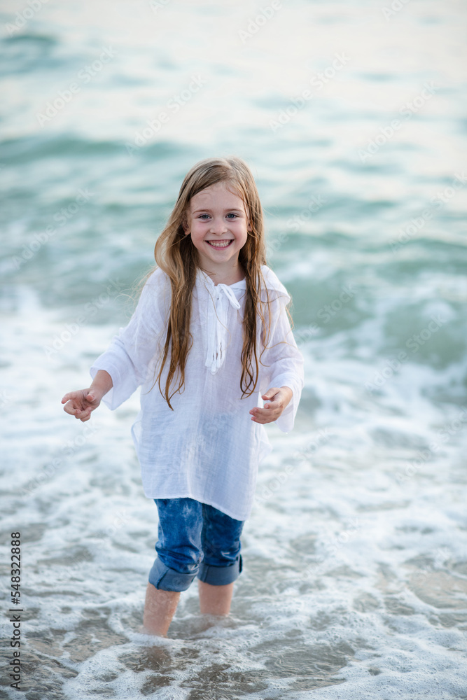 Smiling funny kid girl 5-6 year old wear white top shirt and denim pants walk in sea shore beach outdoor. Childhood. Summer season. Smiling funny kid girl 5-6 year old wear white top shirt and denim pants walk in sea shore beach outdoor. Childhood. Summer season.