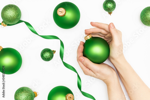 Flat lay of female hands holding green christmas ball with green ribbons and christmas balls on the white background