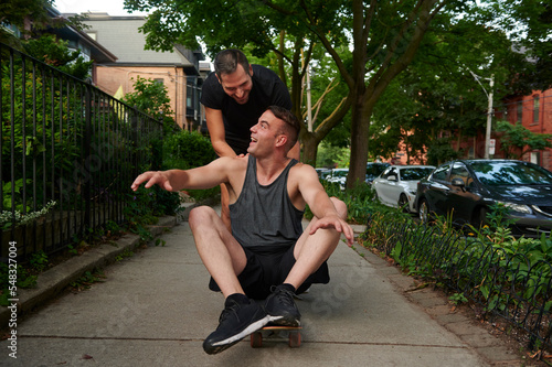 Male pushing same sex partner on skateboard.