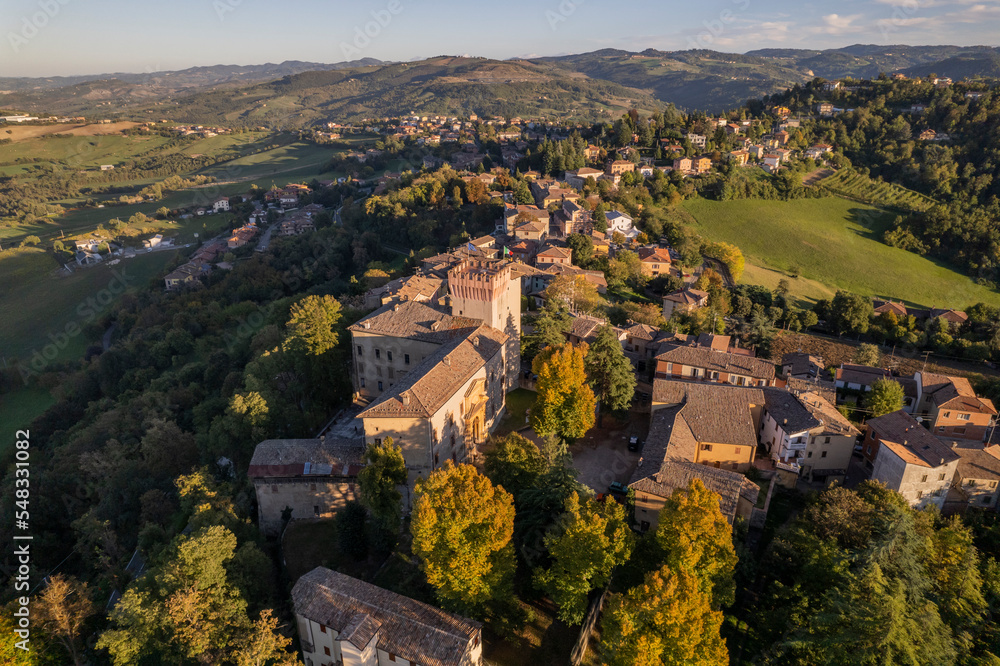 Aerial lateral view of Castello di Guglia among green fields and trees with orange leaves during Fall season in Emilia Romagna in Italy
