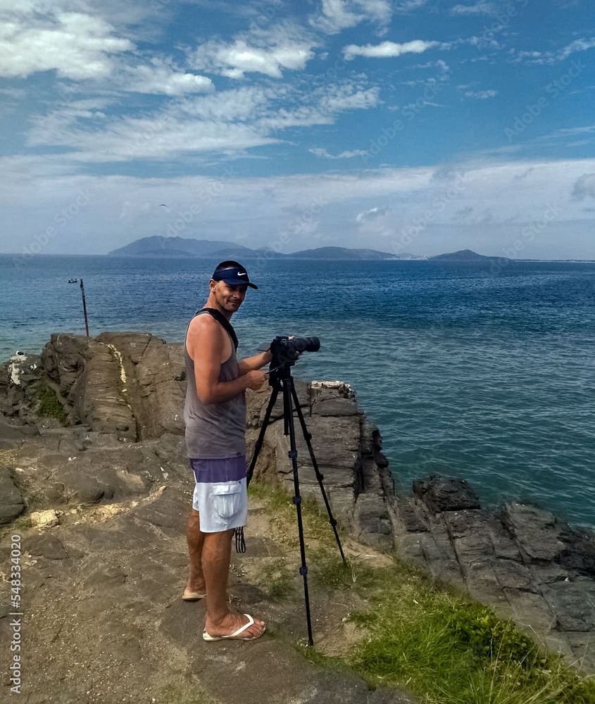Homem tirando foto de cima de rocha na Praia do Forte, com céu azul e linda vista em volta.