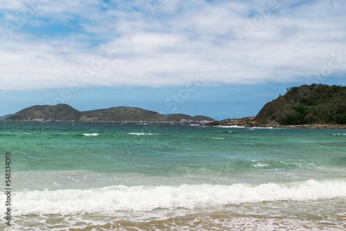 Linda praia das Conchas, próxima a cidade de Cabo Frio, com mar de águas verdes, céu com nuvens e montanhas ao fundo.