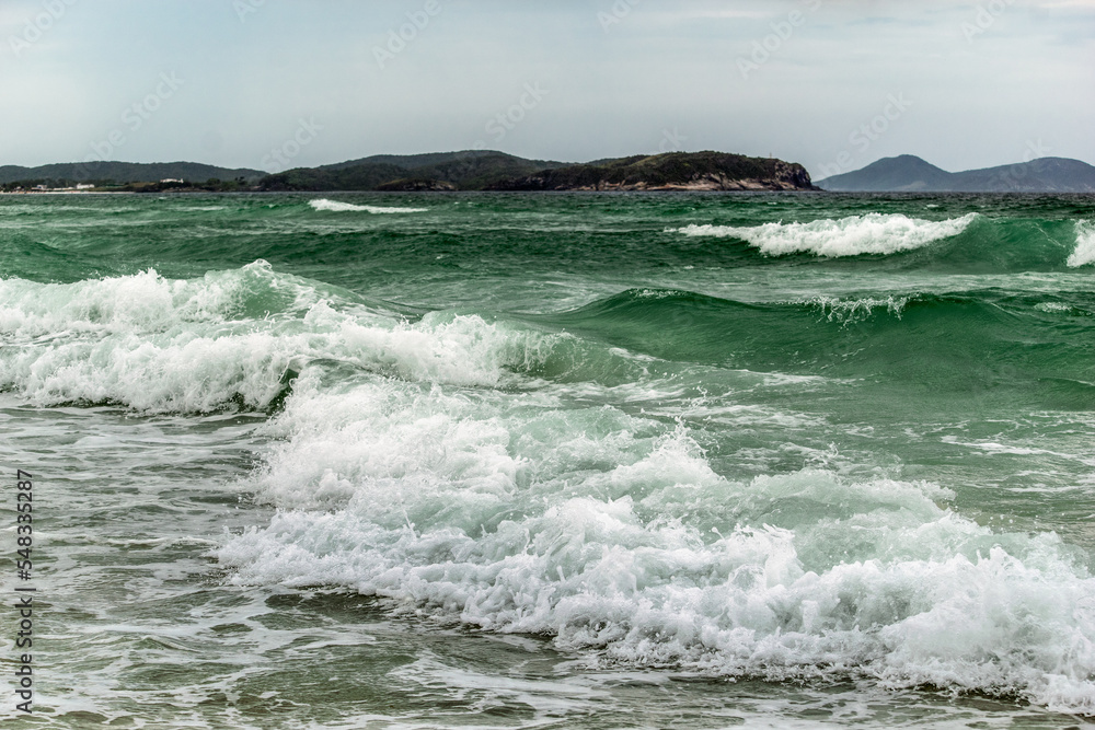 Orla da Praia das Dunas, com um lindo mar, fortes ondas, céu azul e muitas montanhas ao fundo.