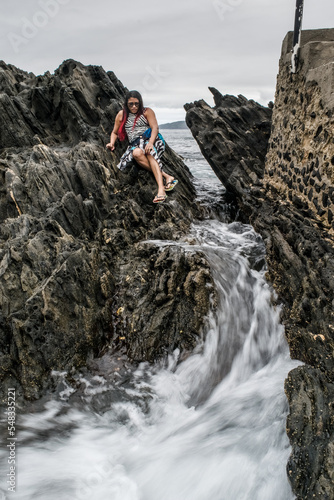 Mulher sentada em rochas onde a água do mar entra nelas formando passagens e possas, praia no Forte São Mateus em Cabo Frio. photo