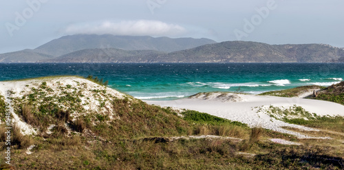 Orla da Praia das Dunas  com algumas dunas de areia  vegeta    o rasteira  c  u azul e muitas montanhas ao fundo.