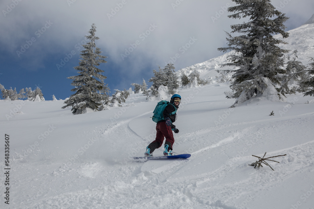 An active female freeriding on a snowboard in a backcountry alpine terrain among snow-covered spruces in white mountains