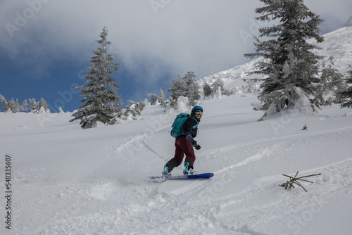 An active female freeriding on a snowboard in a backcountry alpine terrain among snow-covered spruces in white mountains