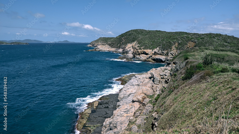 Parte da linda praia das Conchas, com vegetação rasteira, um lindo mar azul em volta, muitas rochas, céu azul e montanhas ao fundo.