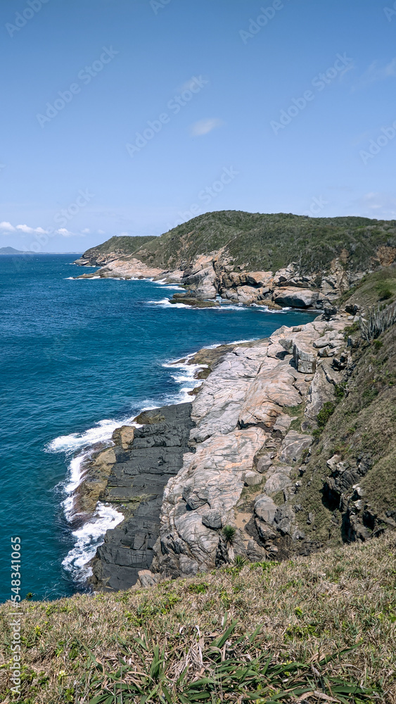 Vista de parte da linda praia das Conchas, com vegetação rasteira, um lindo mar azul em volta, grandes rochas, céu azul e montanhas ao fundo.