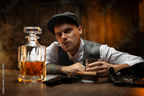 Bold gangster sitting with cigarette at table with cards, pistol and decanter of whisky