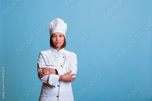 Asian smiling chef standing with arm crossed in studio with blue background preparing culinary recipe using healthy ingredients. Woman cook with white uniform and apron cooking meal dish.