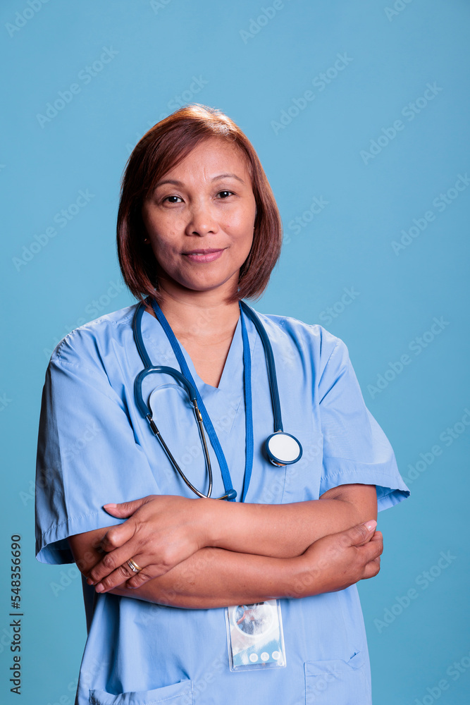 Smiling physician nurse wearing stethoscope and blue uniform during medical consultation working in health care industry. Asian assistant smiling at camera standing in studio with blue background Smiling physician nurse wearing stethoscope and blue uniform during medical consultation working in health care industry. Asian assistant smiling at camera standing in studio with blue background