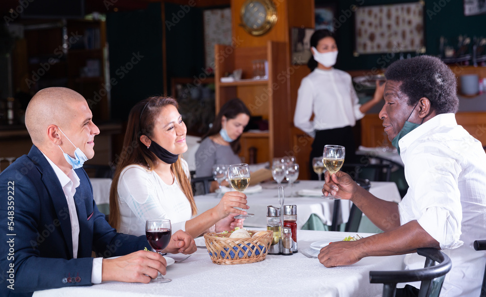 Cheerful people in protective masks having friendly meeting at dinner in restaurant, talking and drinking wine. Concept of reopening restaurants after quarantine due to coronavirus