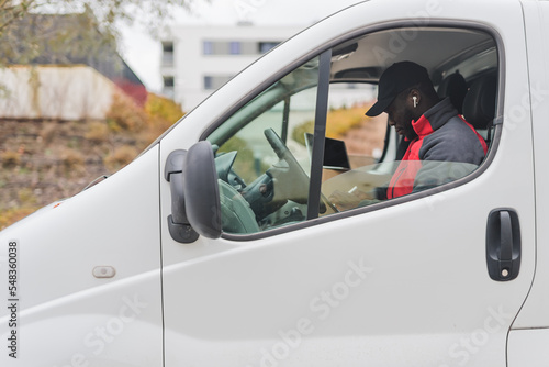Hard-working, focused ethnic male courier during his shift. African-American man in black baseball hat sitting behind the wheel of white delivery van and checking things on his clipboard. High quality