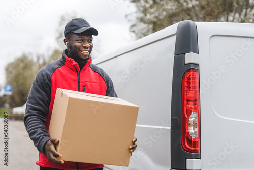Package delivery concept. Outdoor medium closeup shot of middle-aged African-American handsome deliveryman in red jacket holding a big package and standing in front of white delivery truck. High