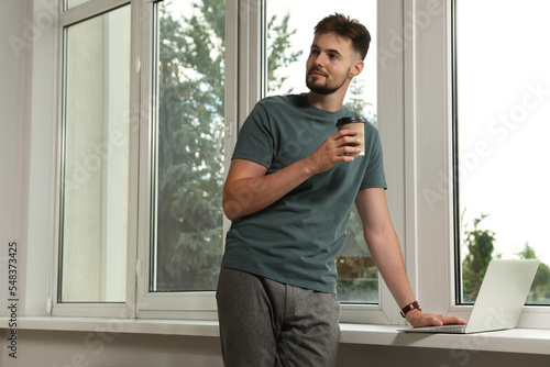Handsome man with laptop and cup of coffee near window indoors, space for text