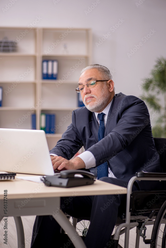 Old male employee in wheel-chair sitting at workplace Old male employee in wheel-chair sitting at workplace